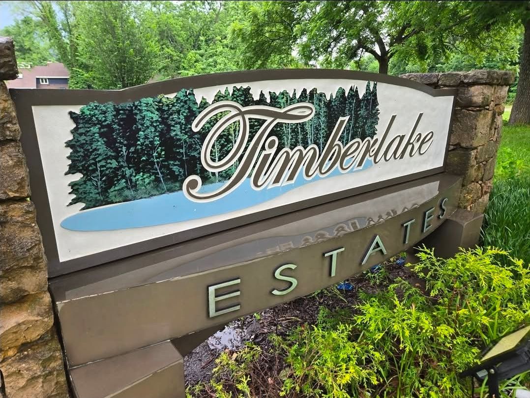 A stone monument sign for Timberlake Estates with a graphic of a forest and lake, set in a green landscaped area.