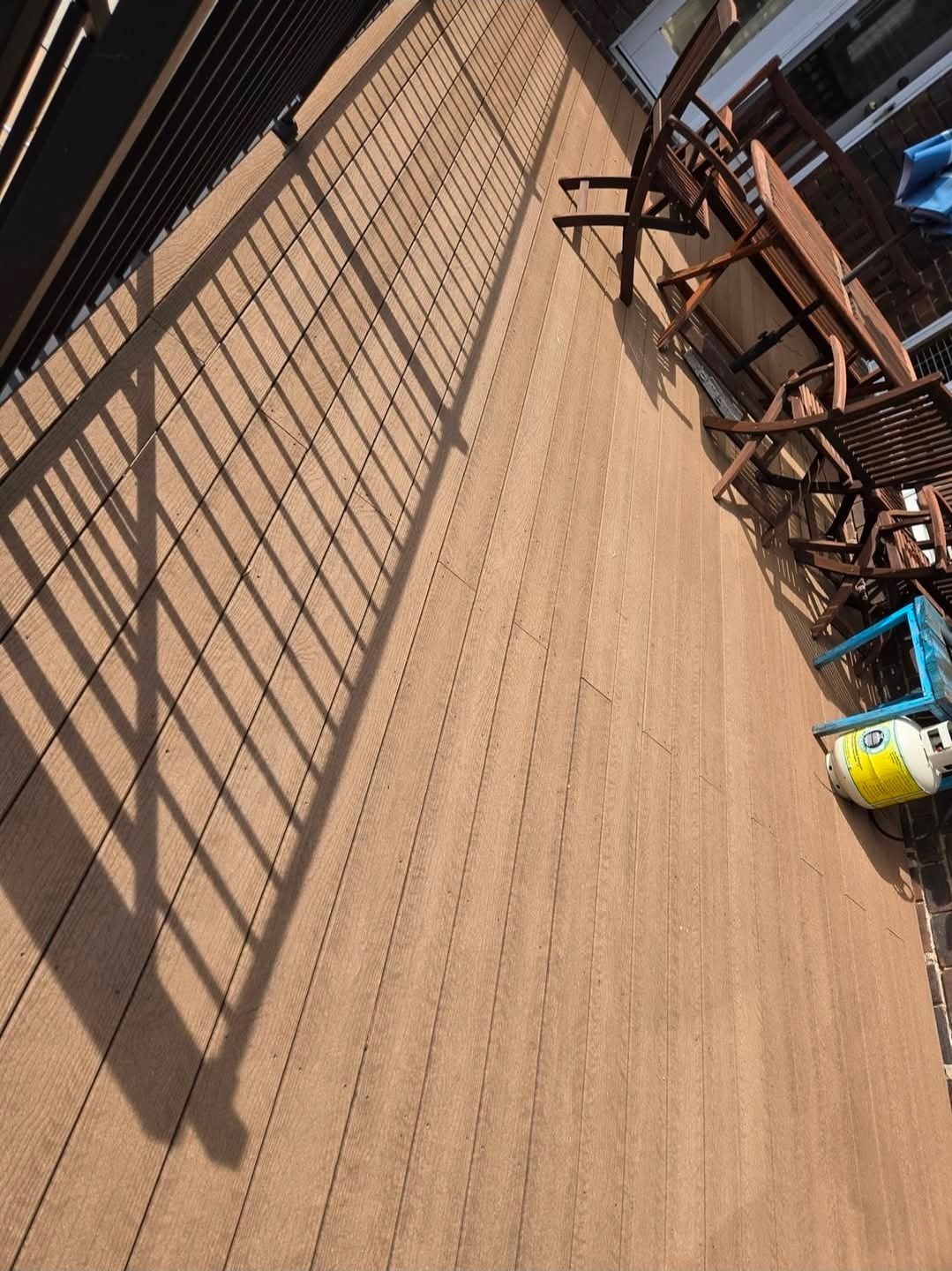 A wooden deck featuring a dining table, chairs, and a propane tank in the corner, viewed from above with a railing shadow.