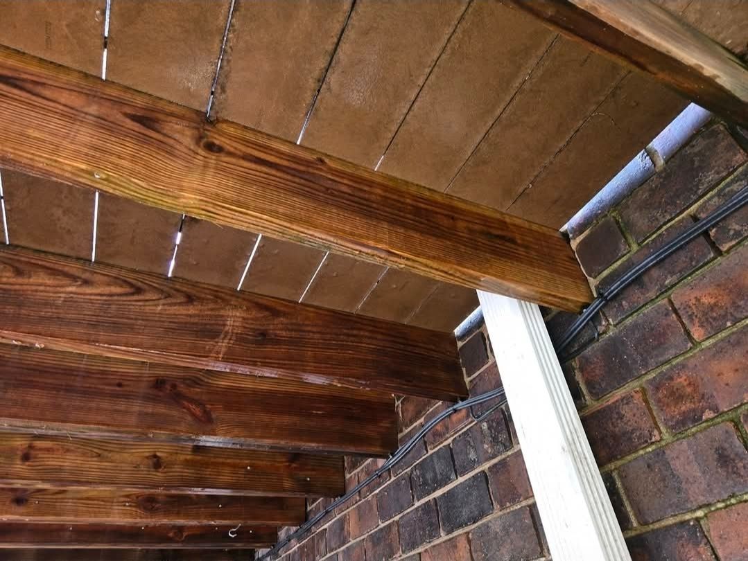 A low-angle view showing dark stained wooden beams supporting a deck, adjacent to a brick wall with a white downspout.