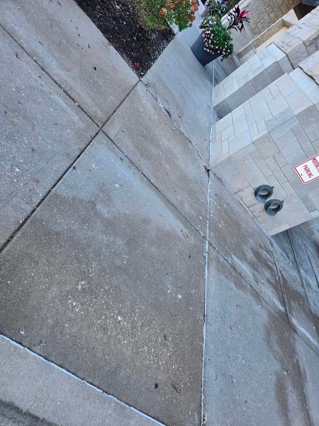 A concrete sidewalk corner with a crack along the foundation of a brick building near a small potted plant.