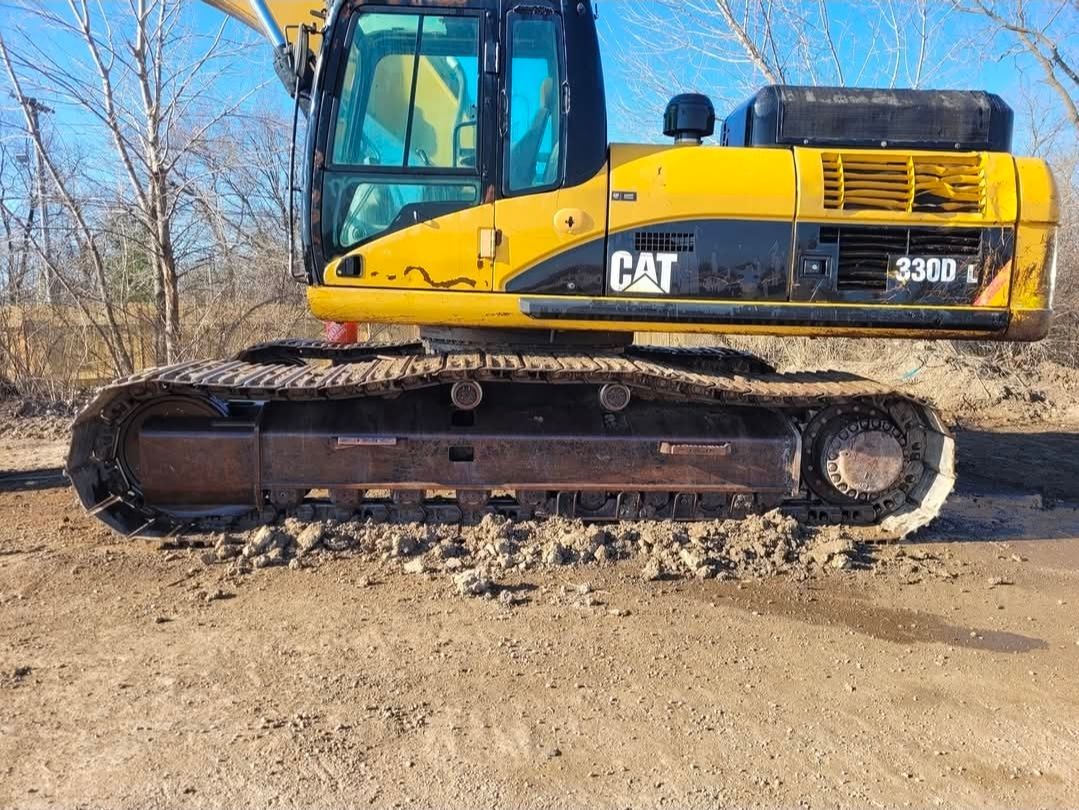 A yellow Caterpillar 330D L hydraulic excavator sits on dirt ground with a treeline in the background.