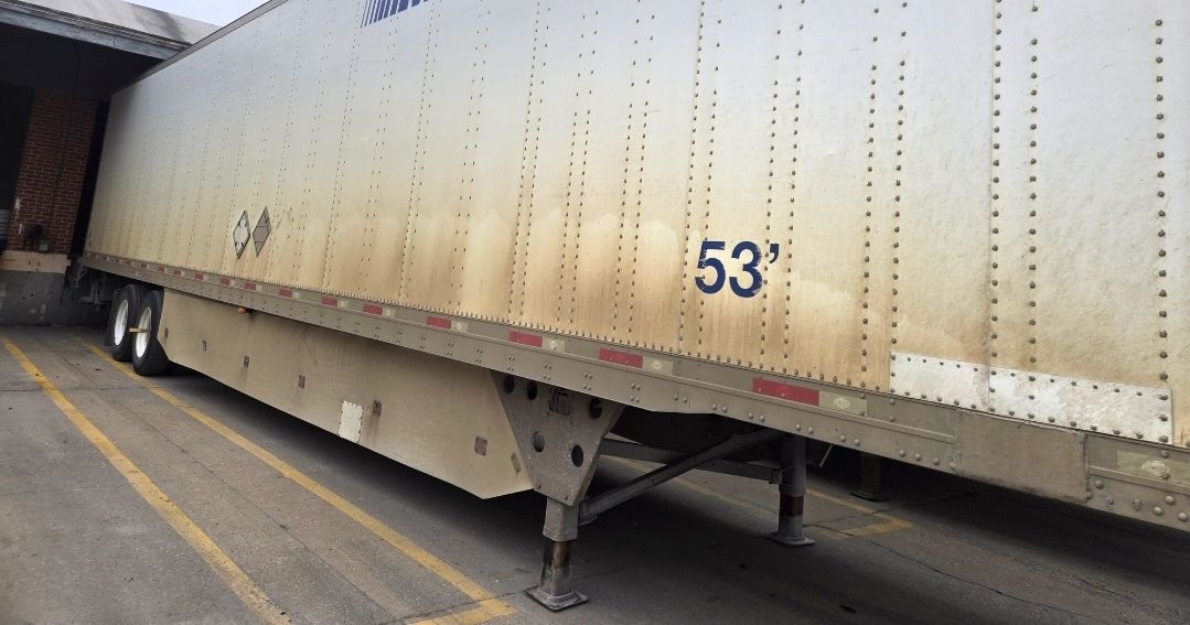 A side view of a dirty, beige 53-foot semi-trailer docked at a warehouse loading bay.