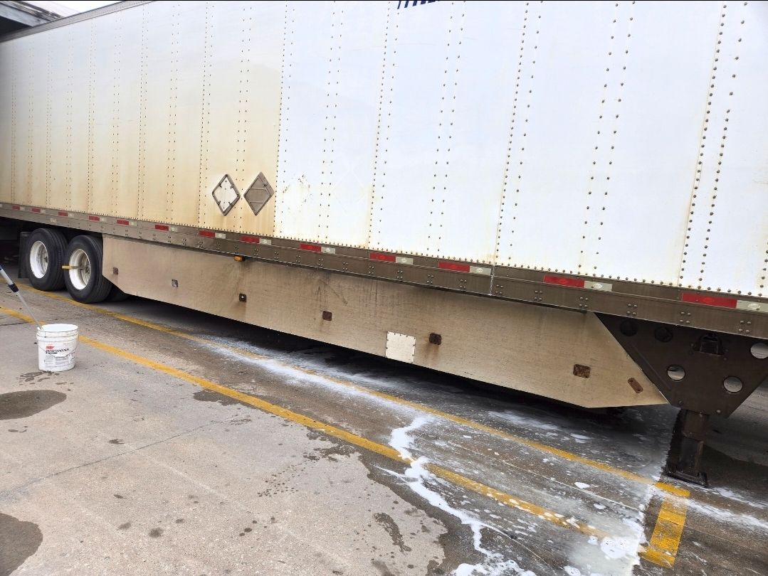 A white trailer parked on concrete with a white foamy substance pooling underneath it next to a bucket.