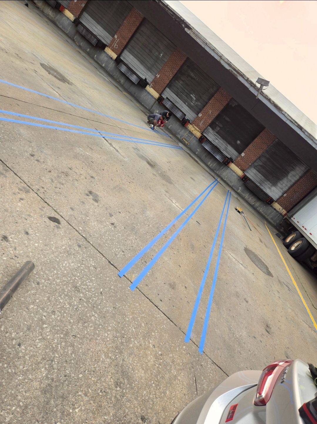 A person kneels near a row of loading docks at a concrete building, with blue-painted parking lines on the ground.