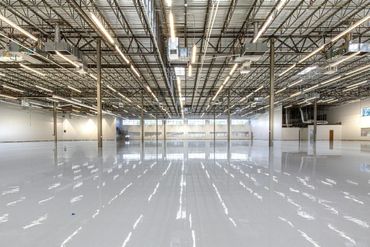 A vast, empty warehouse with white polished concrete floors and exposed industrial ceiling beams with bright lighting.