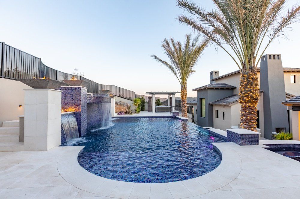 A large swimming pool with a waterfall and palm trees in front of a house.