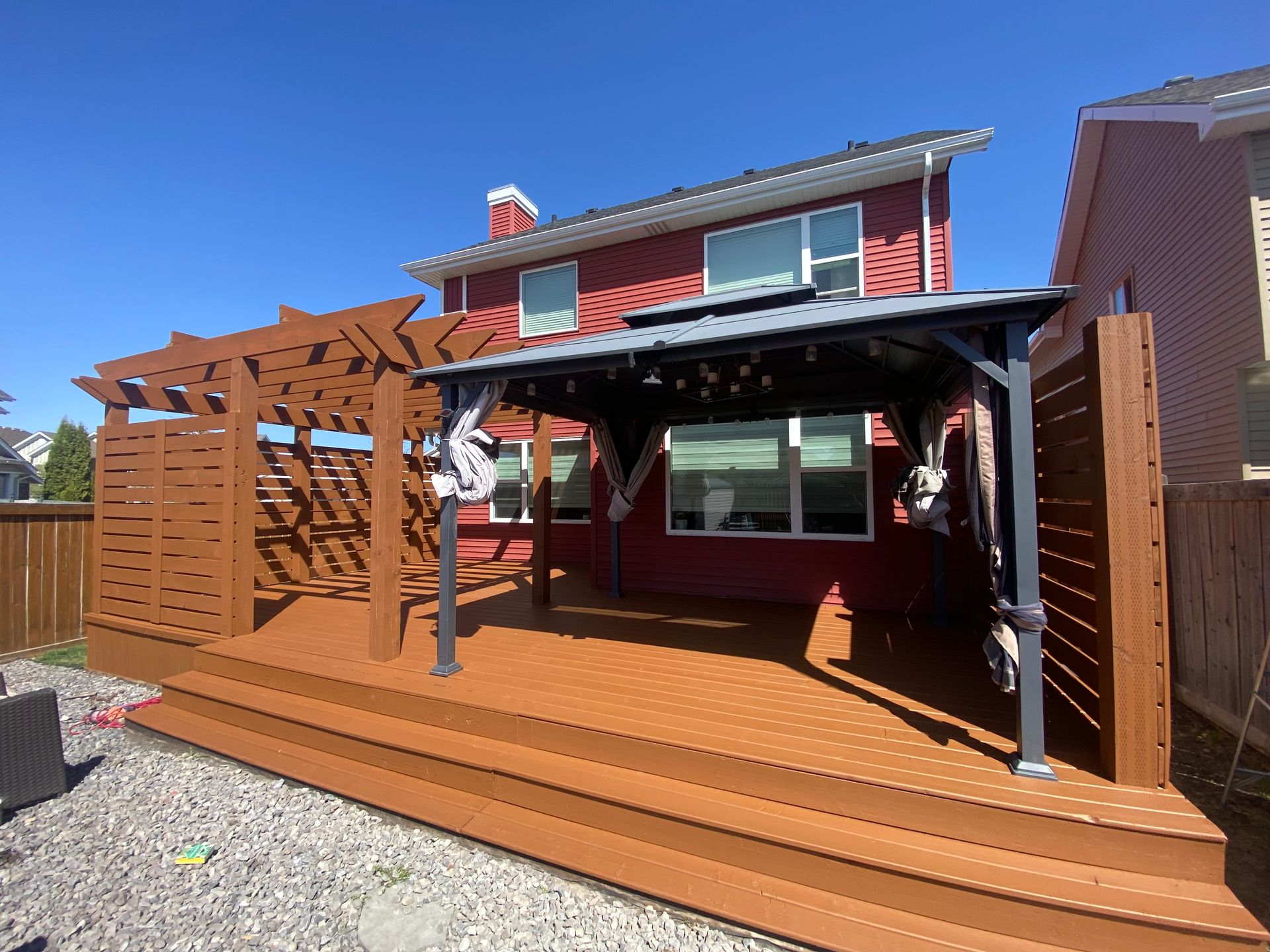 Wooden deck with pergola and red house under a blue sky.