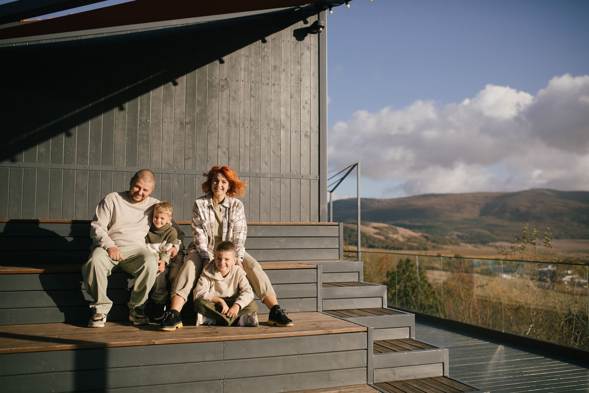 Three people sitting on a wooden deck by a cabin, with mountains and clouds in the background.