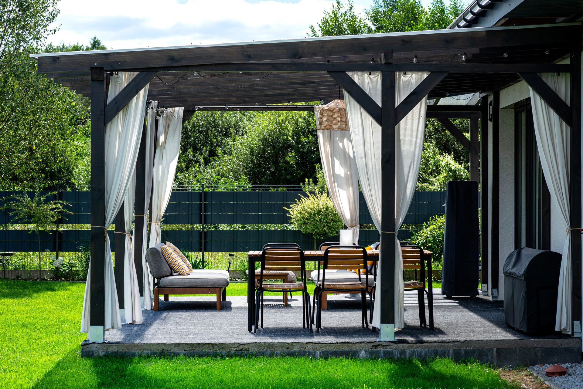 Covered patio deck with white curtains, wicker chairs, and a wooden table on a sunny lawn