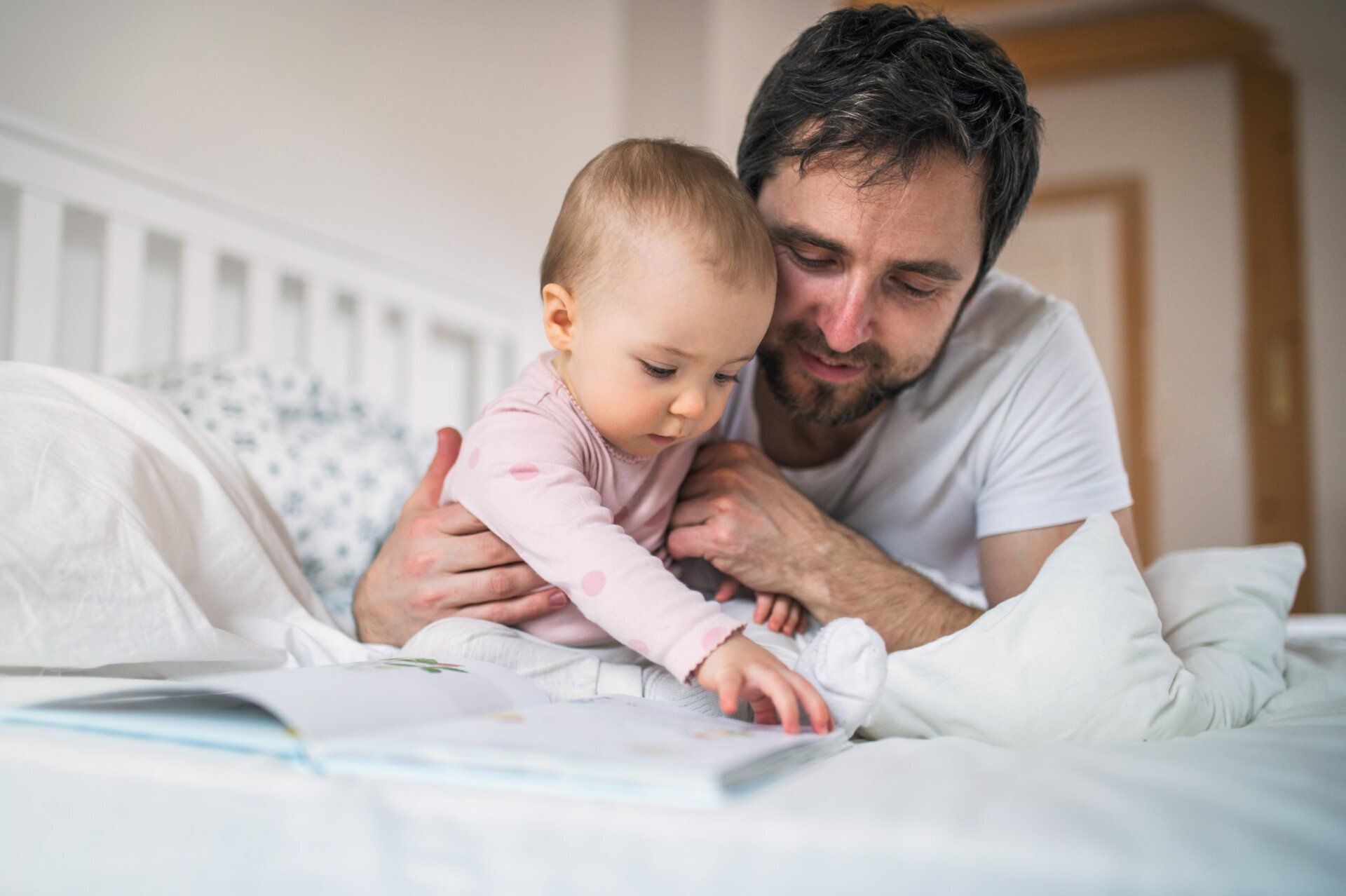 Adult holding baby on a bed, reading together in a bright bedroom