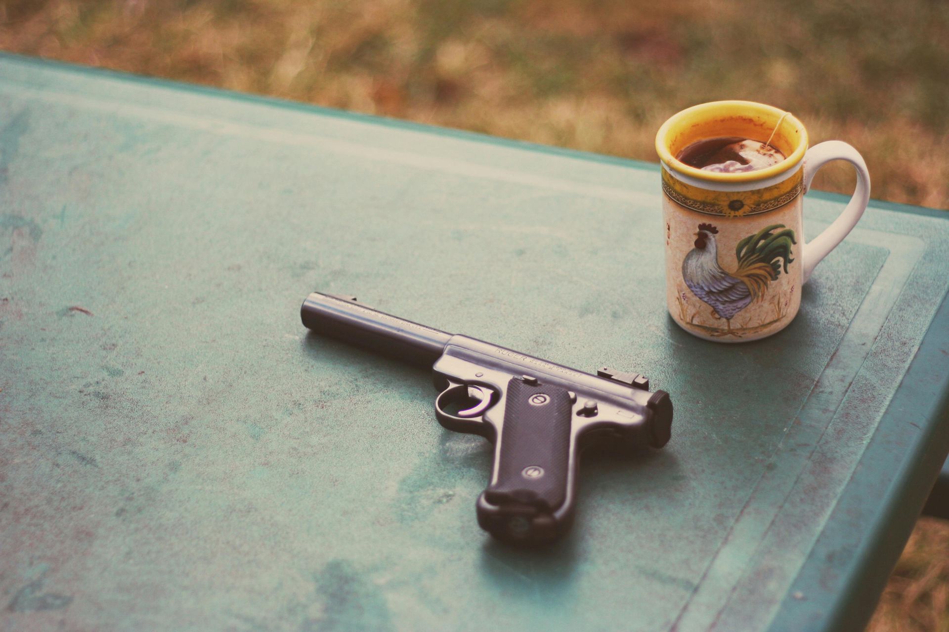 Gun and mug of coffee on a green table outdoors. Mug has rooster design.