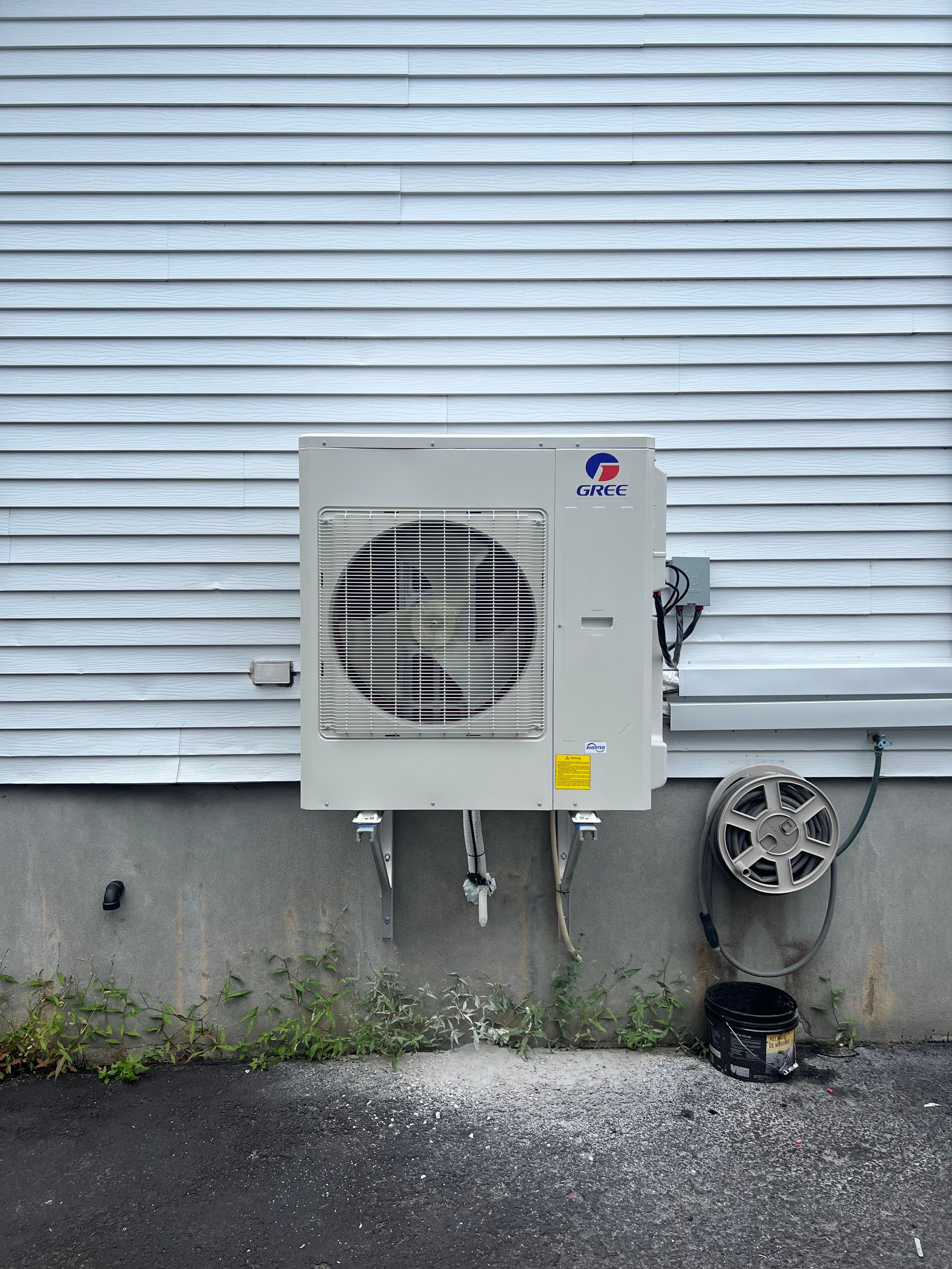 Une unité de climatisation blanche fixée sur un mur bardé gris au-dessus de fondations en béton, avec des débris au sol en dessous.