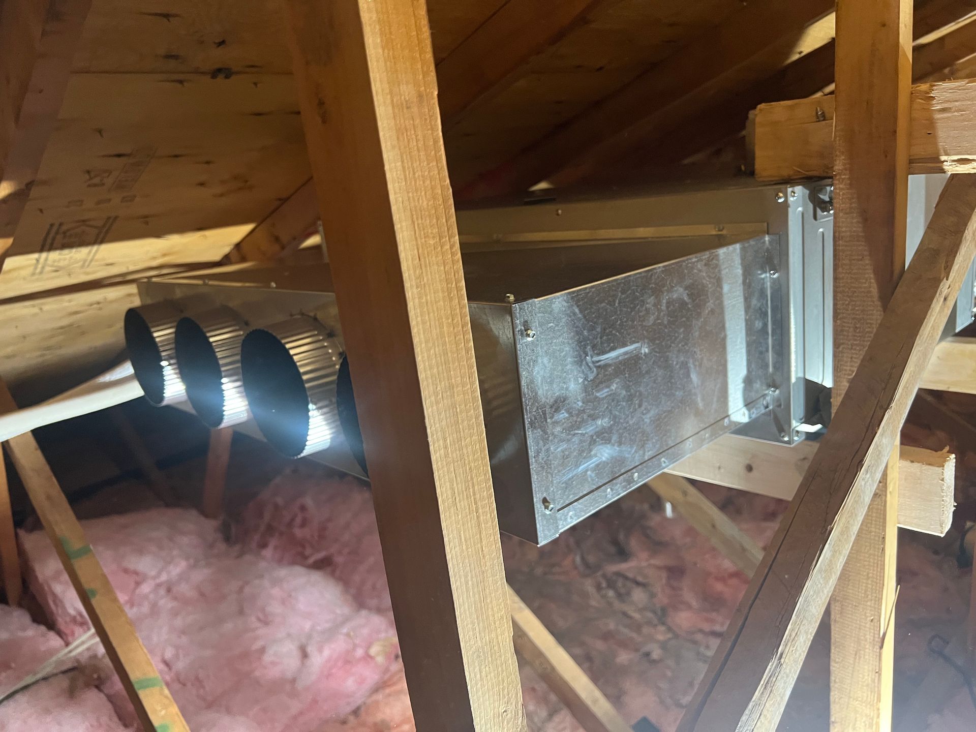 Conduits de ventilation métalliques avec plusieurs orifices circulaires suspendus entre les chevrons en bois du grenier, au-dessus d'une isolation rose.