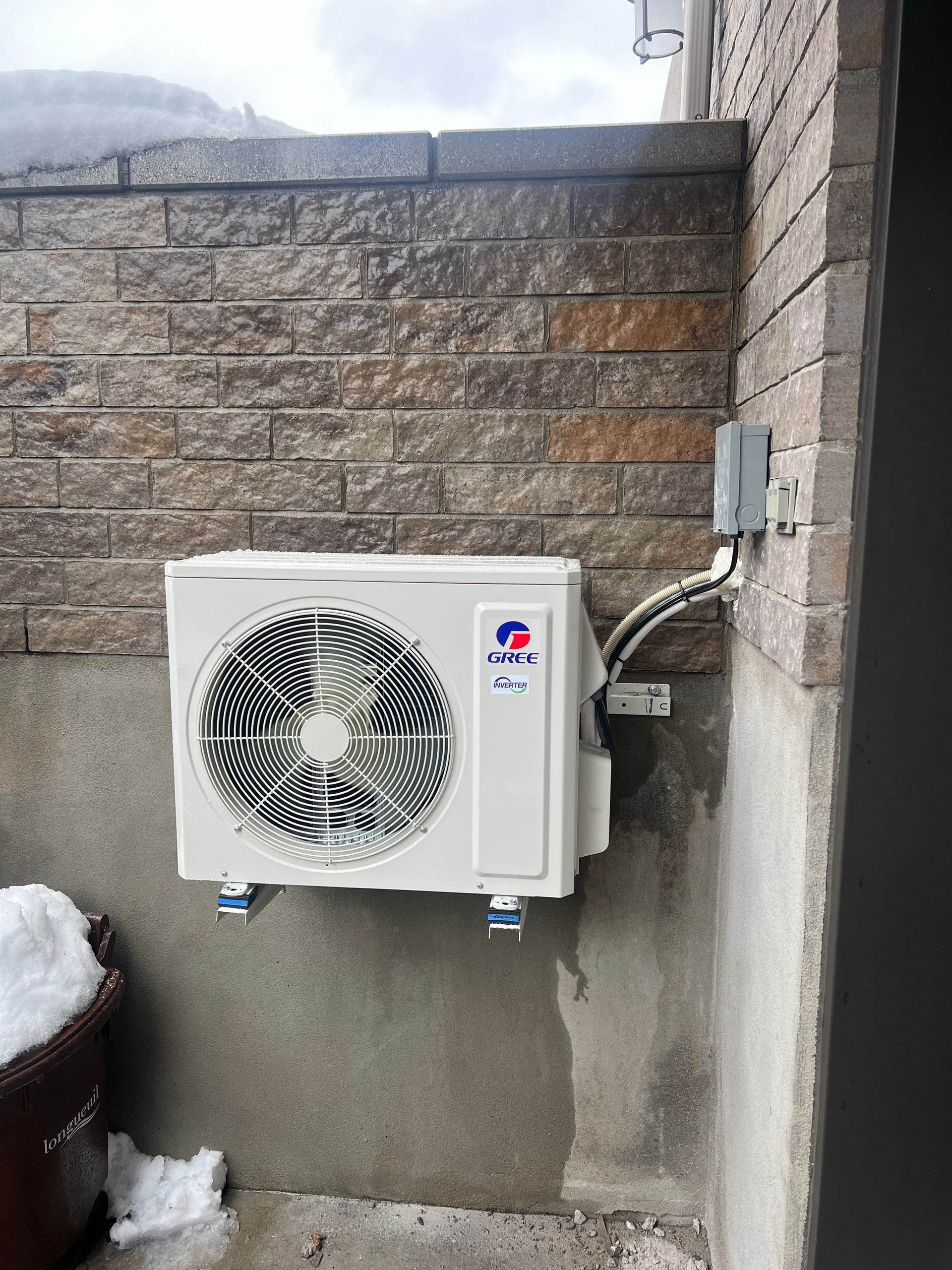 Une pompe à chaleur Gree blanche, montée sur un mur en pierre et en béton, avec un boîtier de déconnexion électrique à droite.