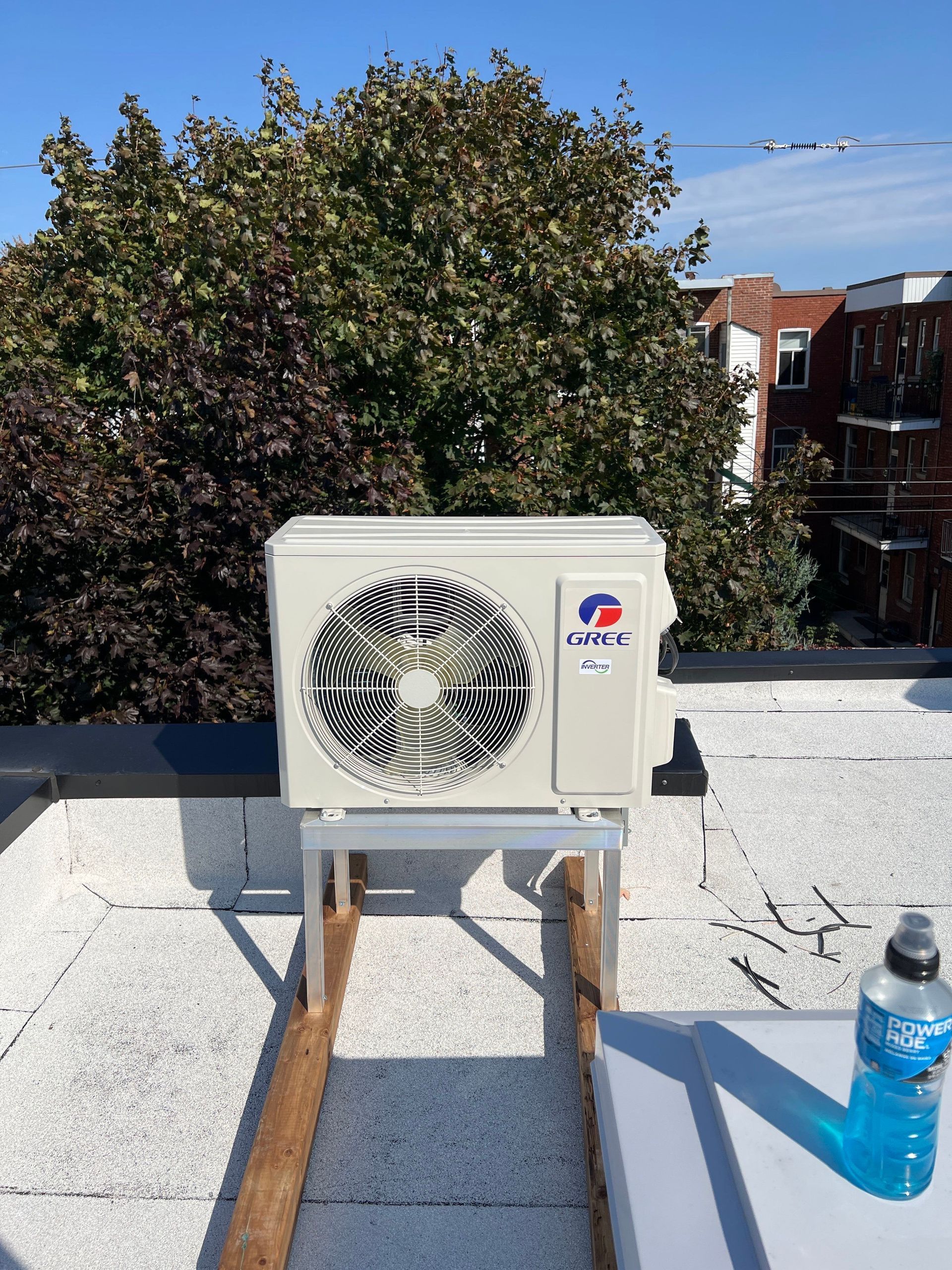 Un climatiseur extérieur Gree beige est installé sur un support métallique, sur un toit plat et blanc, entouré d'arbres et de bâtiments.