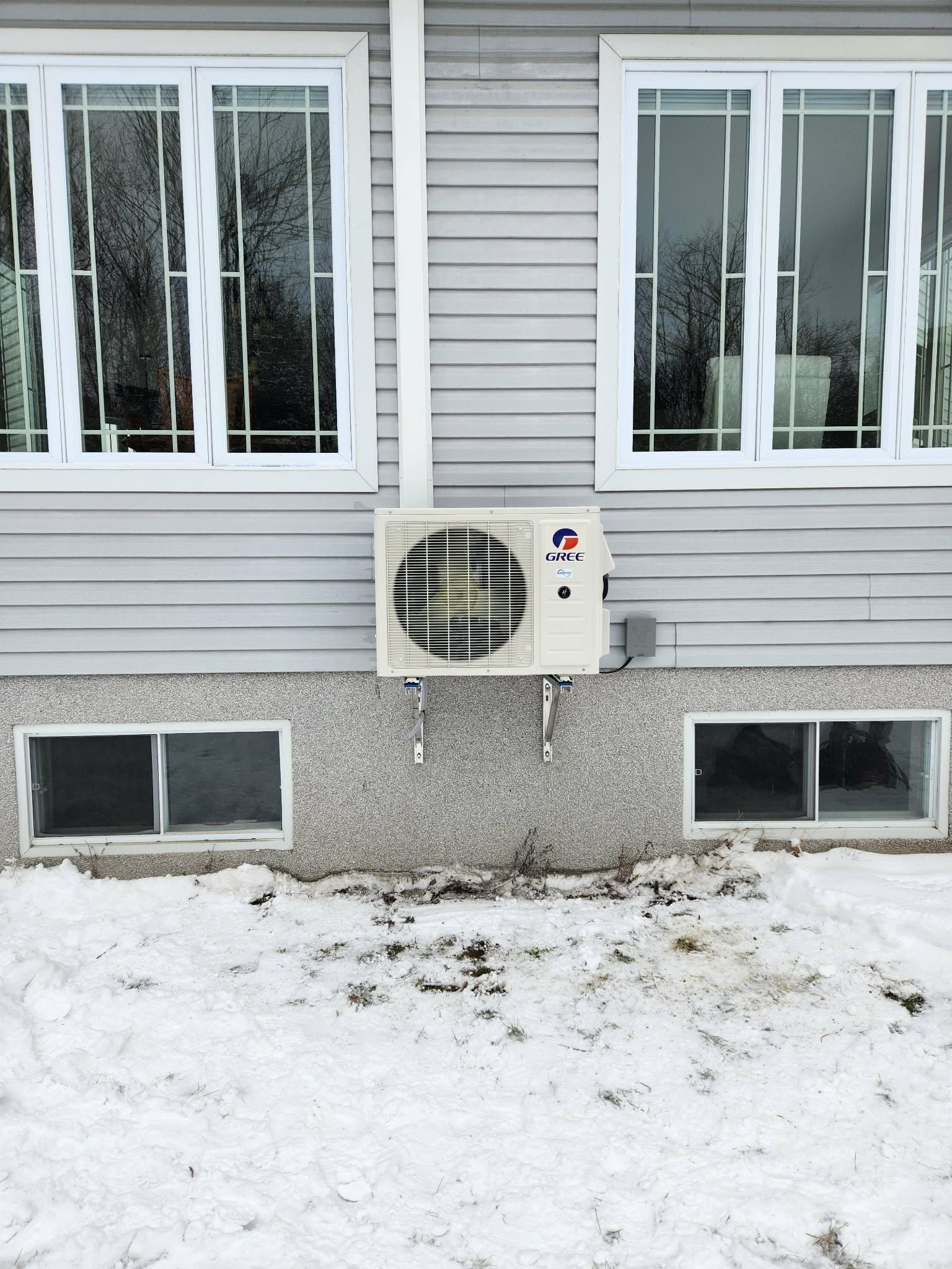Une pompe à chaleur extérieure Gree, montée sur un revêtement gris, située entre deux fenêtres de sous-sol au niveau du sol, sous la neige.