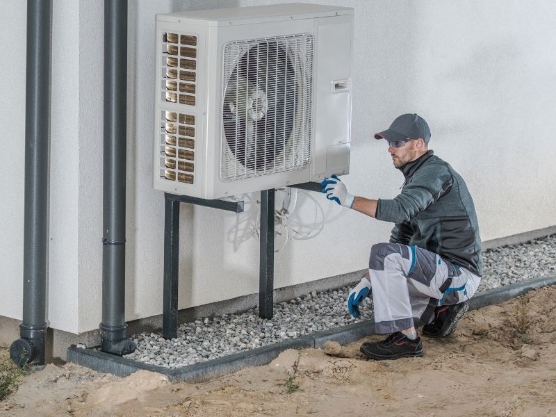Un technicien en tenue de travail et gants s'agenouille pour entretenir une unité de pompe à chaleur de climatisation extérieure, adossée au mur d'une maison.