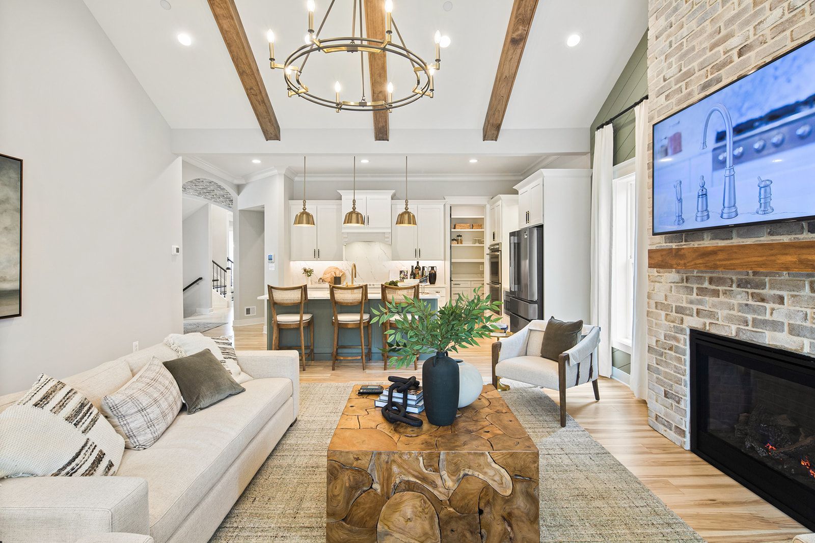 Living room with white sofa, kitchen in background, brick fireplace with TV, and wooden beams.