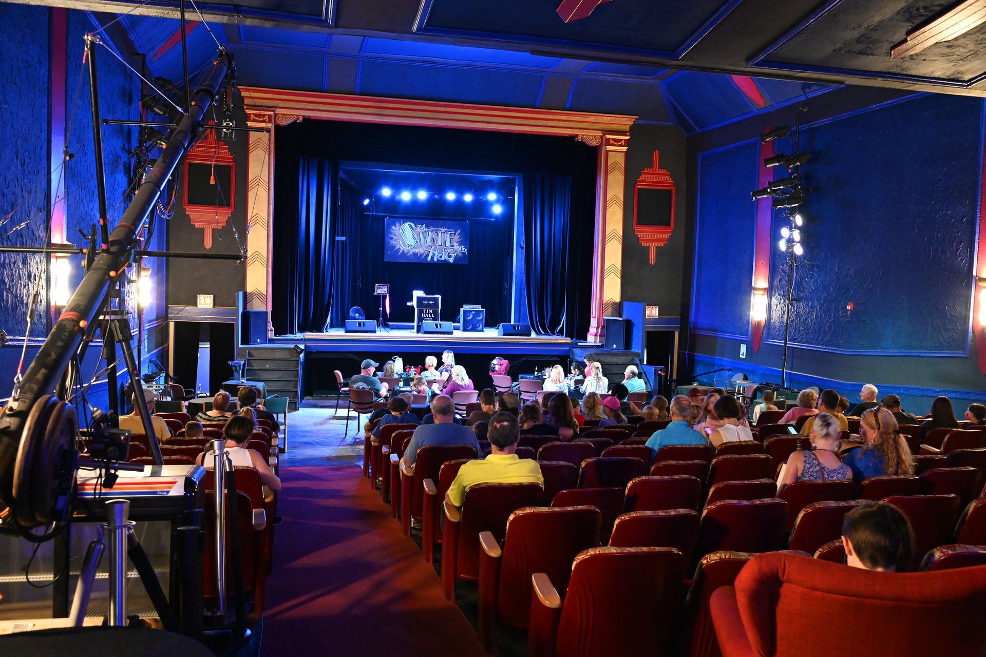 Audience seated in red chairs looking towards a stage with performers in a theater with blue walls.