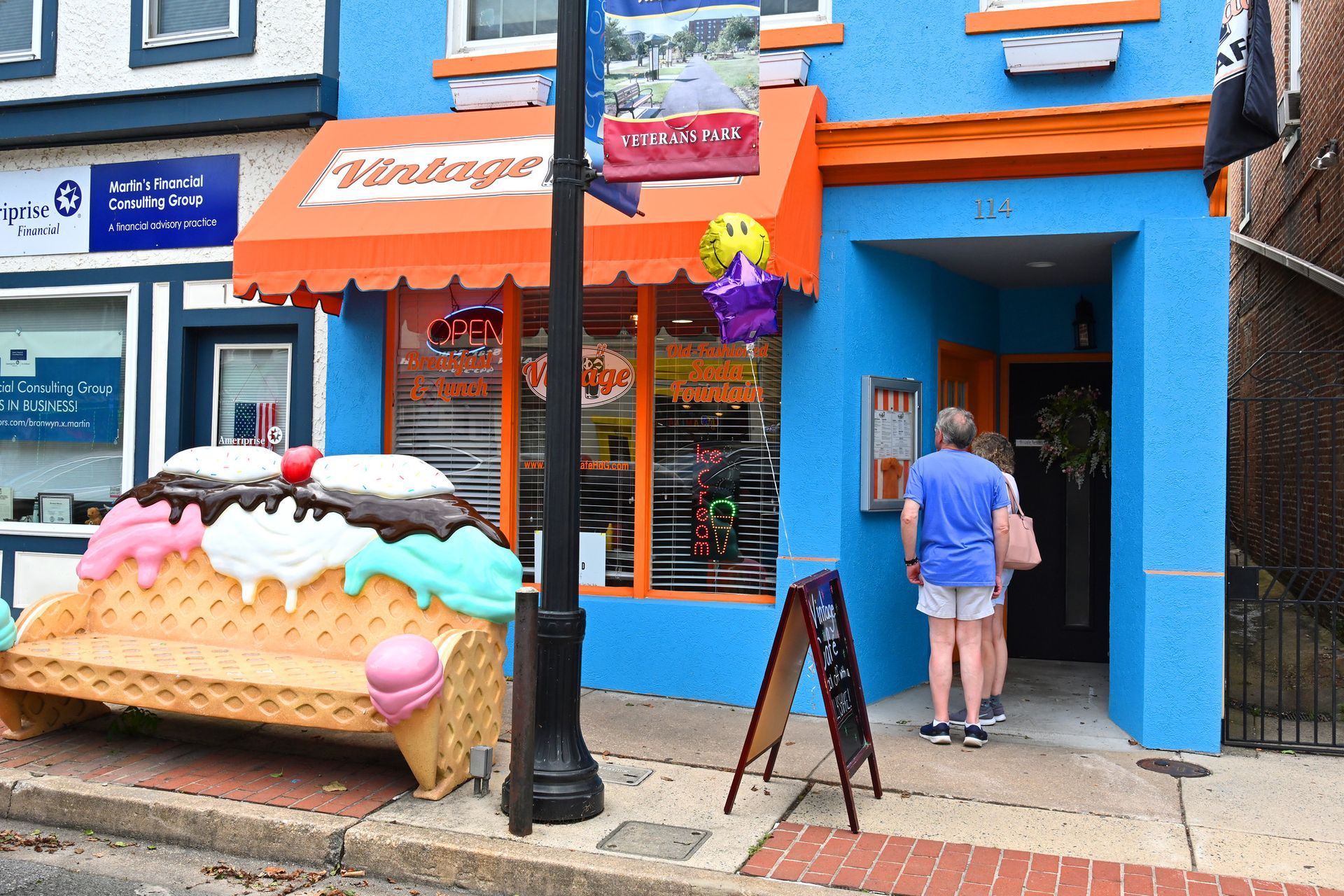 Bright blue ice cream shop with an orange awning. People entering. Giant ice cream bench out front.