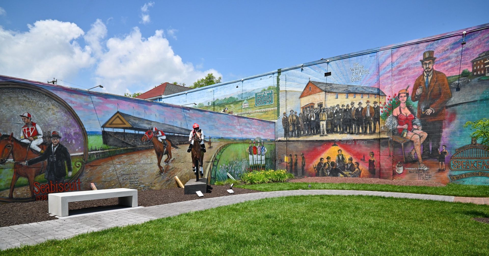 Mural depicting historical scenes with figures on horseback, a crowd, and a woman; bright colors on a brick wall in a park setting.