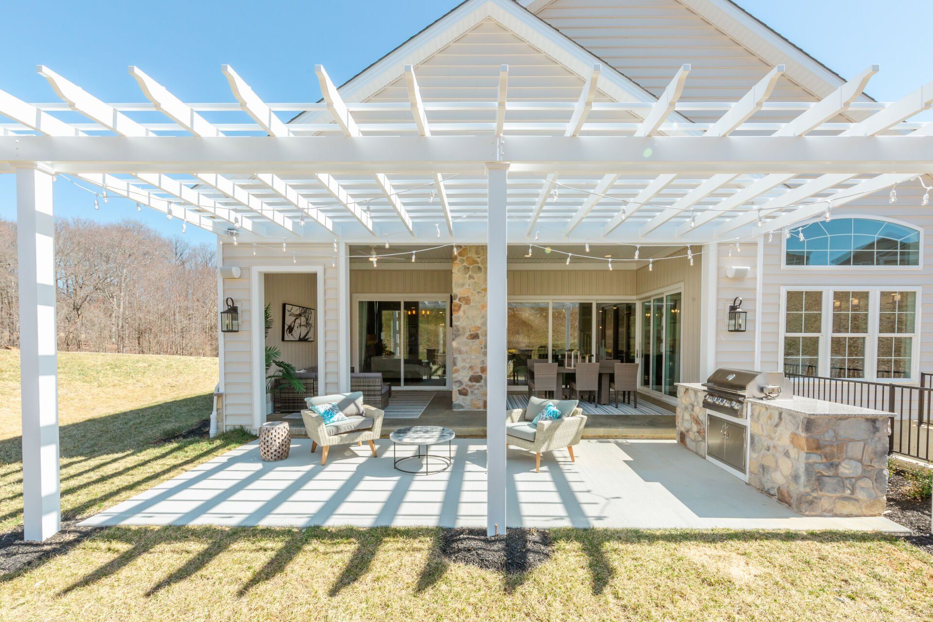 Back patio with pergola, seating area, and grill. Beige siding, string lights, sunny day.