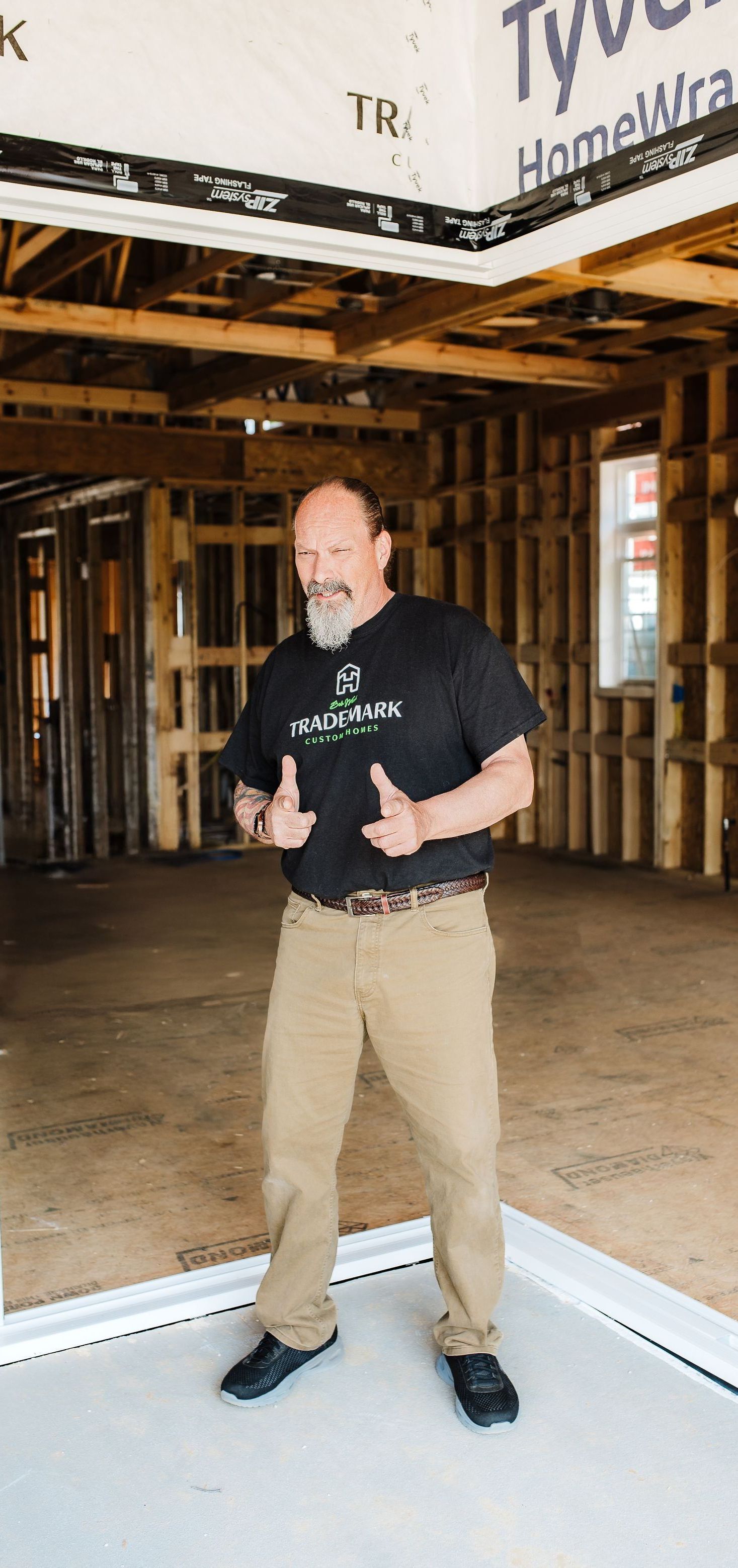 Man standing in an unfinished house, giving a thumbs-up. Wearing a black shirt, tan pants, and black shoes.