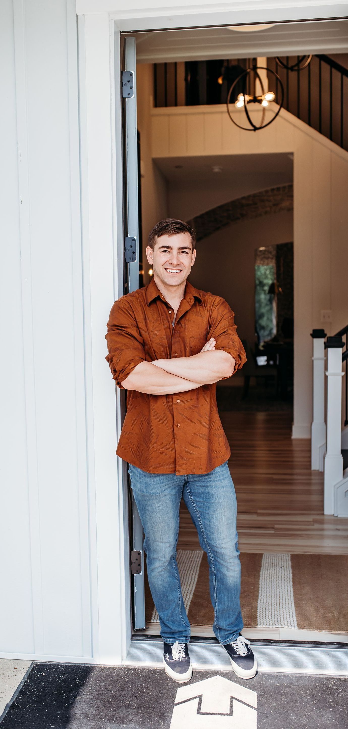 Man in brown shirt and jeans stands in doorway with arms crossed.