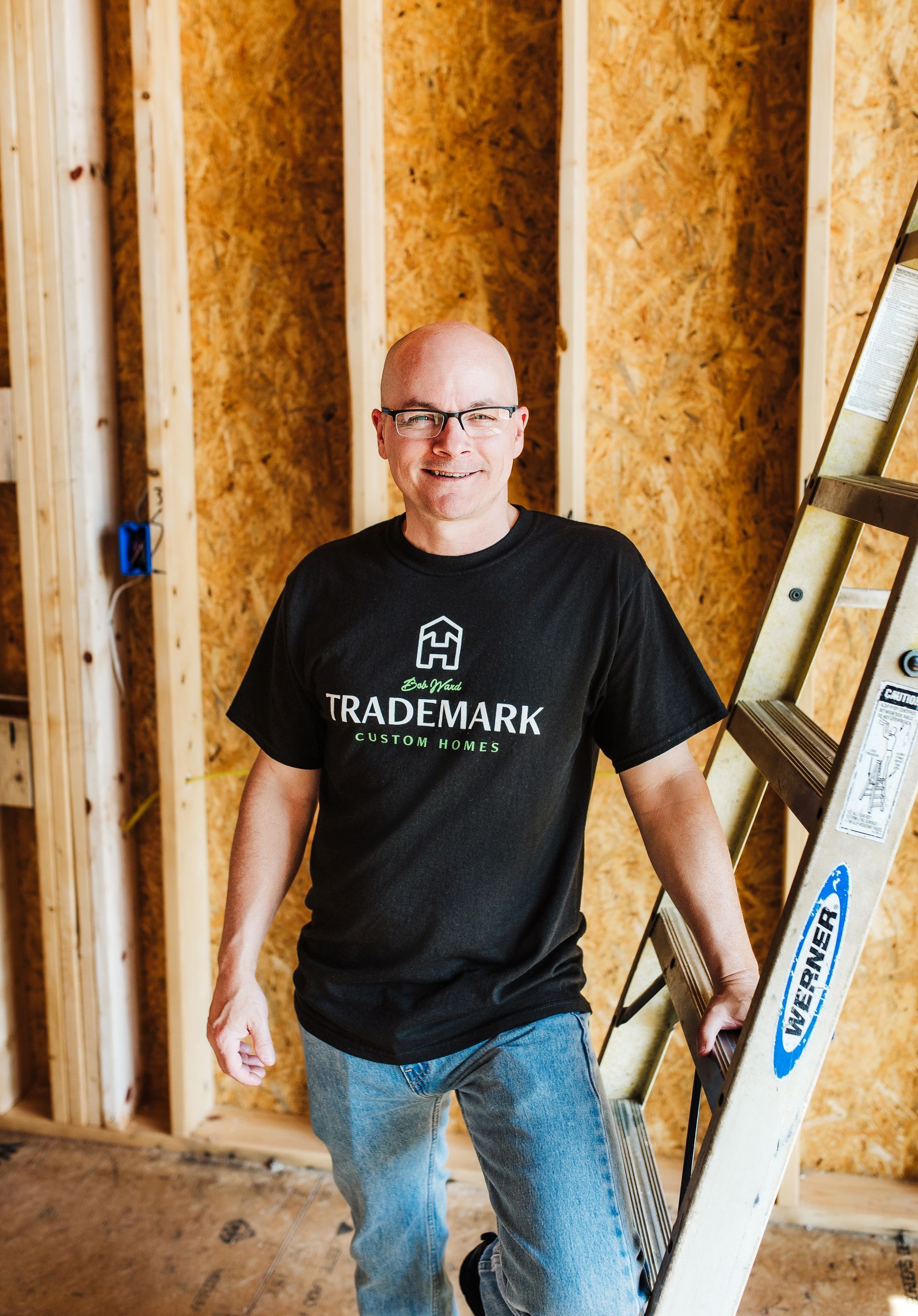 Man with glasses in a black Trademark t-shirt stands in front of wooden wall, next to a ladder.