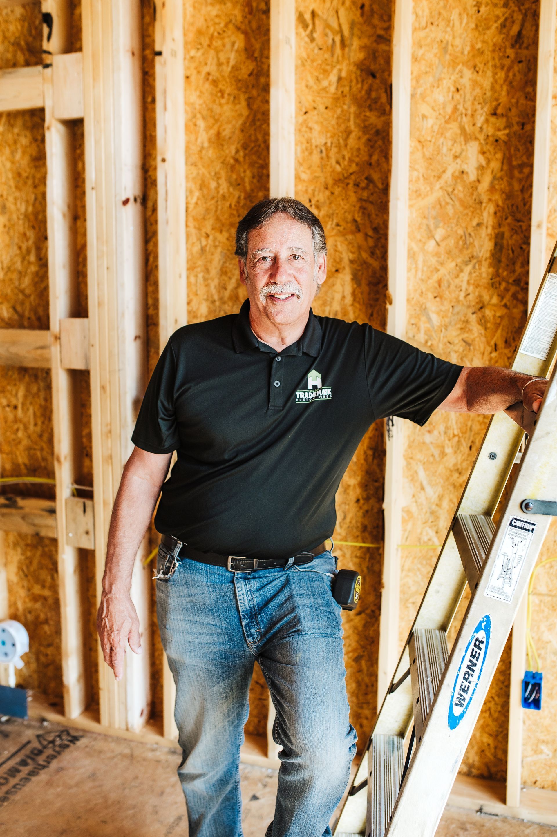 Man in black shirt and jeans leans on a ladder in a construction site.