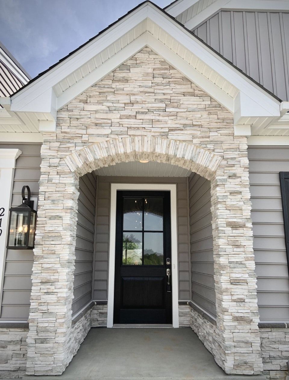 Gray-sided house entrance with stone archway around a black door; outdoor setting.
