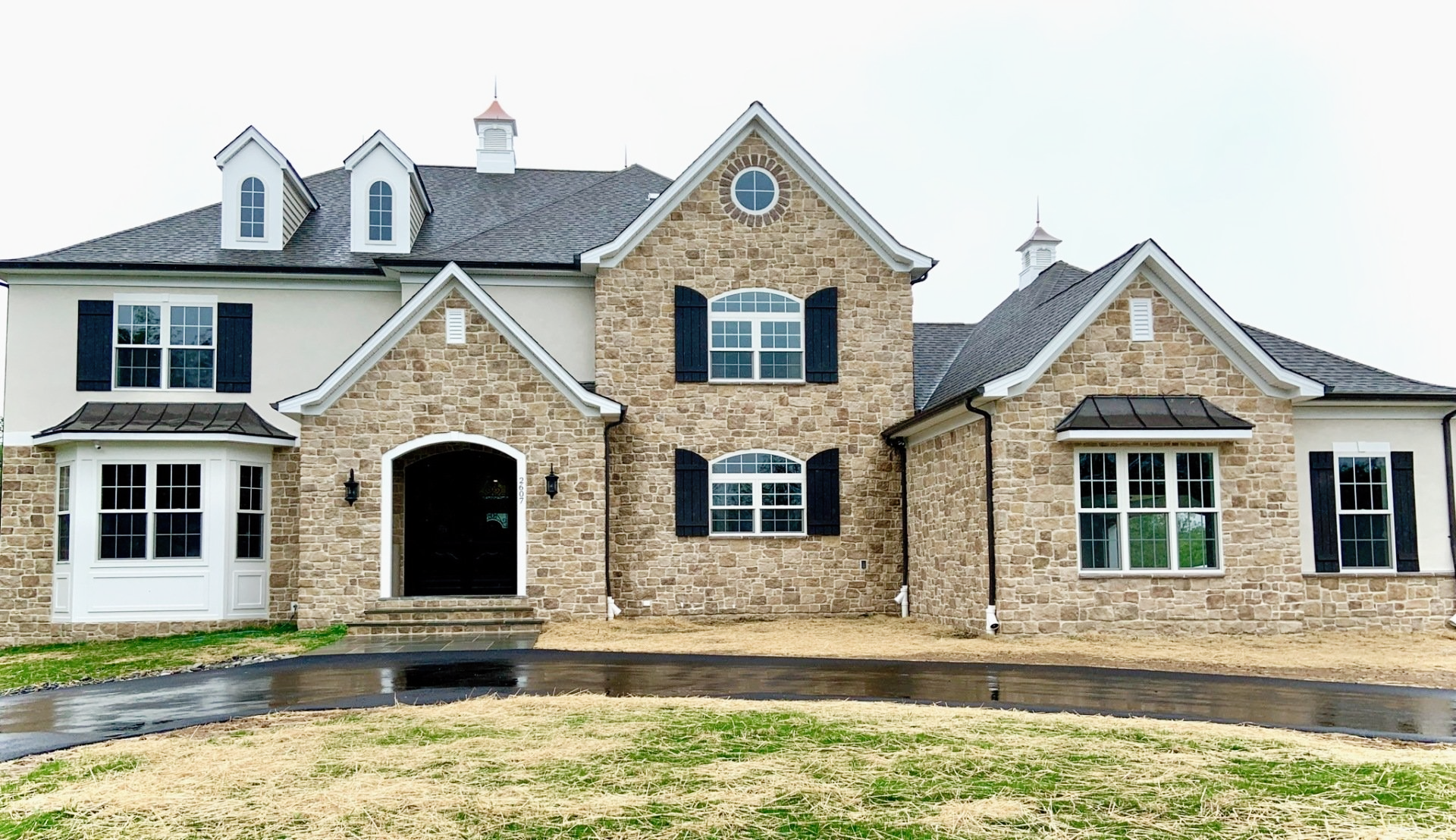 Two-story brick and stucco house with black shutters, dormers, and a wet driveway.