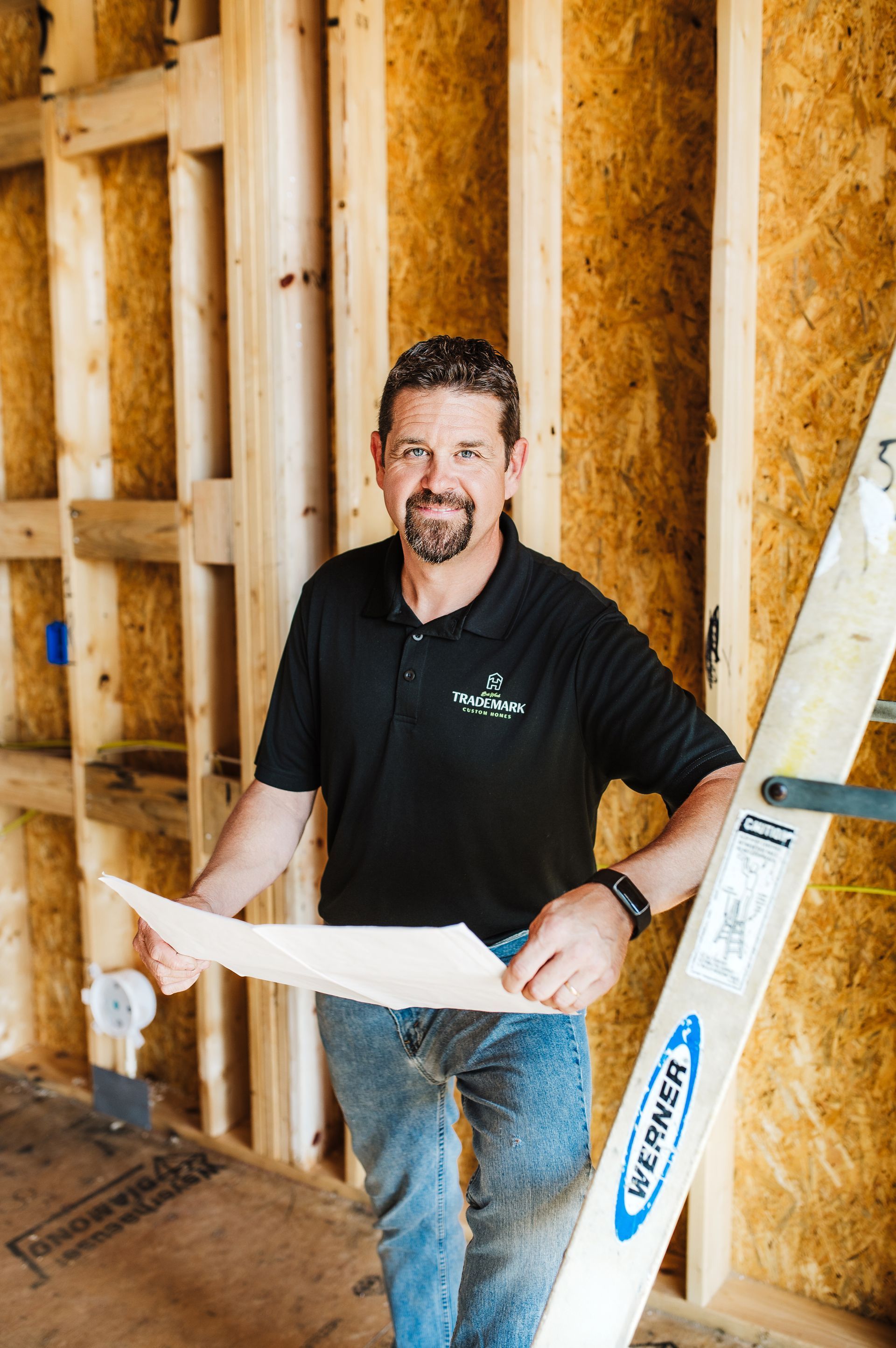 Man in black shirt, holding blueprints, leans against a ladder inside a building frame.