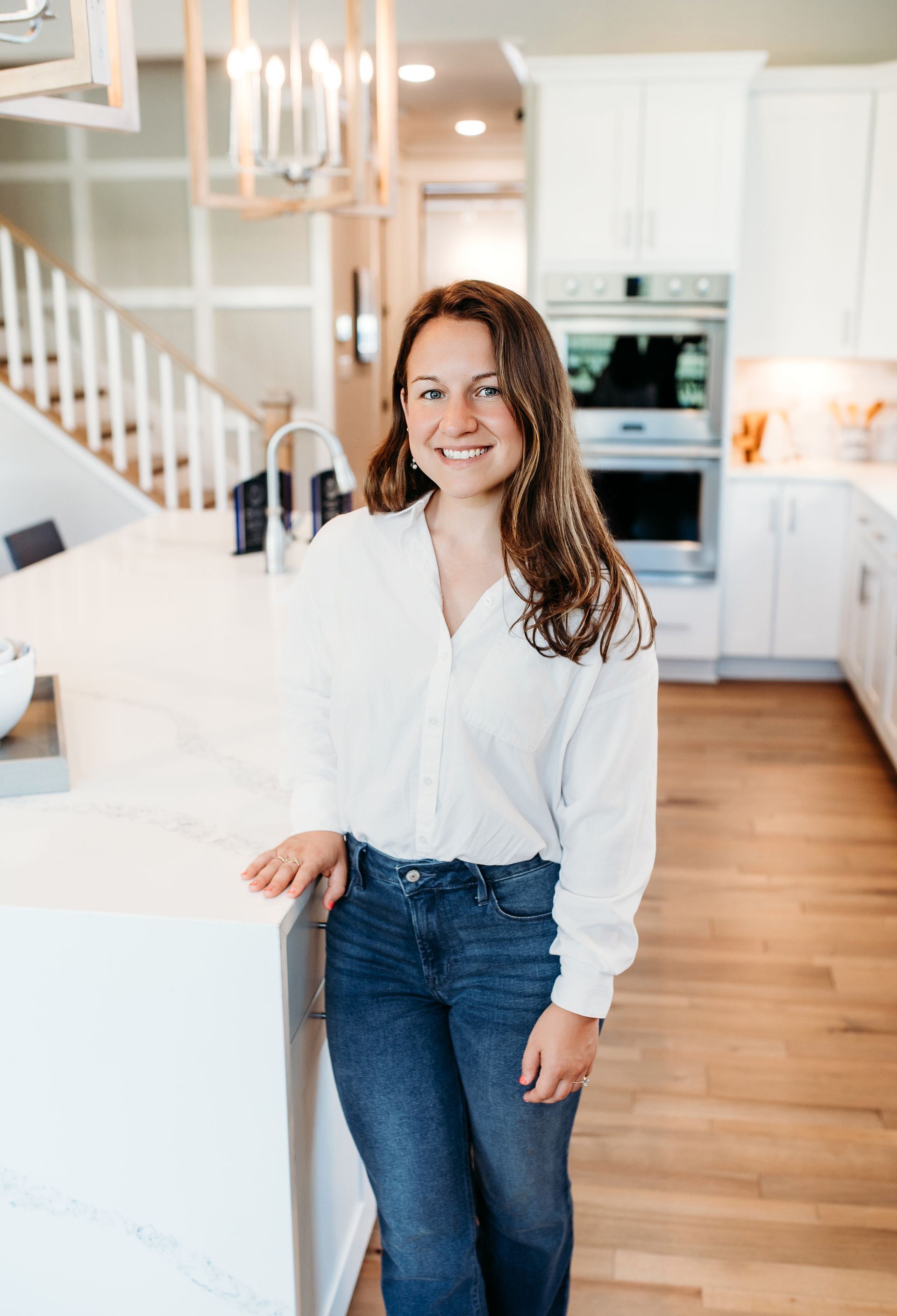 Woman in white shirt and jeans, smiling in a white kitchen, leaning on a white island.