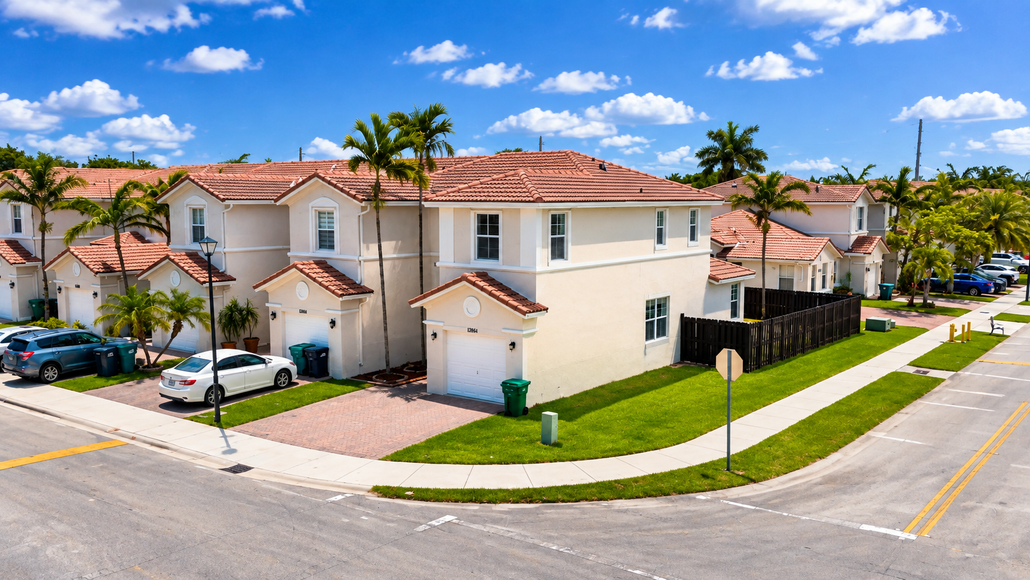 Suburban corner townhouses with red-tile roofs, palm trees, parked cars, and a blue sky