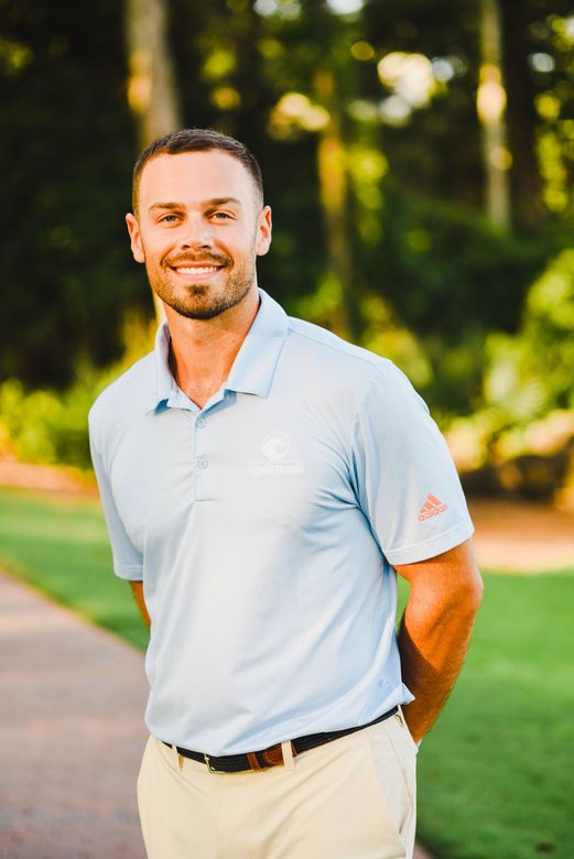 A smiling person in a light blue polo shirt and tan pants stands outdoors in front of a soft-focus, wooded background.