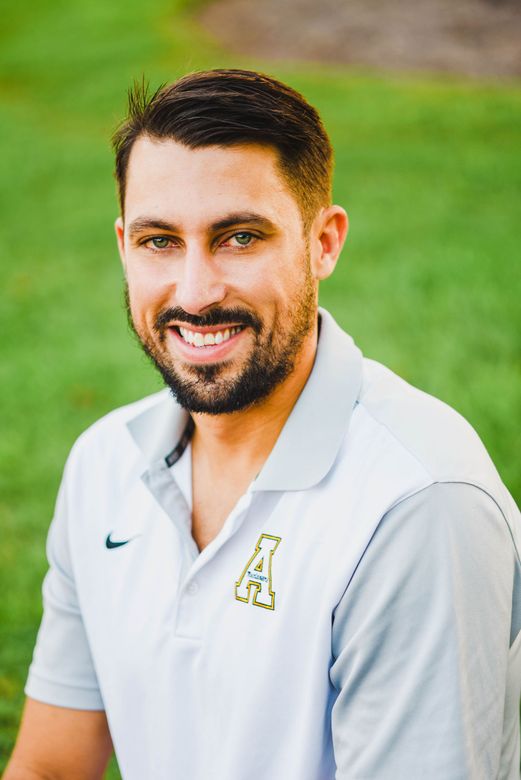 A smiling person with a beard wears a light-colored, short-sleeved polo shirt with a small emblem, posed against green grass.