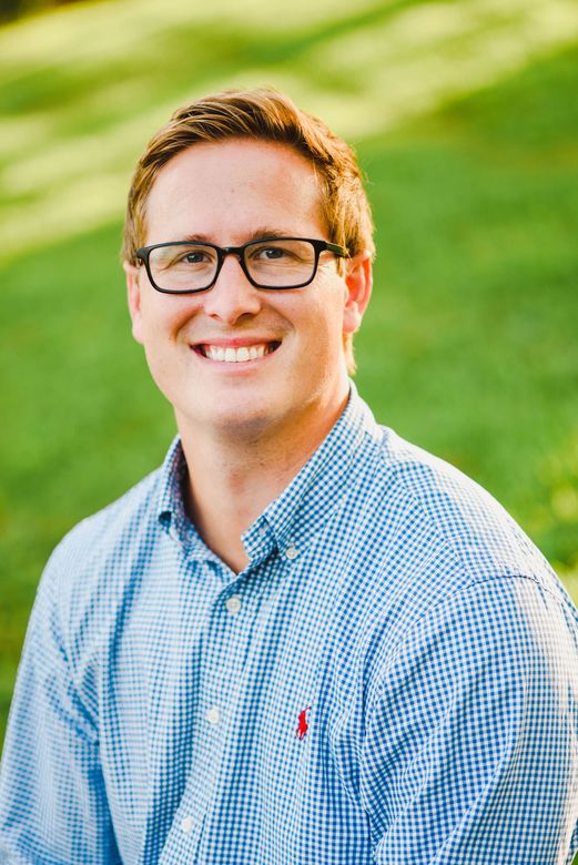 A person with light-colored hair and glasses smiles while wearing a light blue gingham button-down shirt outdoors.
