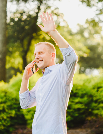 A man in a light blue button-down shirt talks on a mobile phone while waving in a sunlit, leafy outdoor setting.