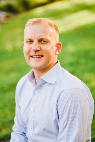 A person wearing a light blue button-down shirt smiles while standing in front of a soft-focus green grassy background.