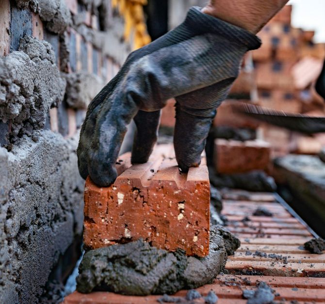 Gloved hand placing a red brick on a mortar bed in a brick wall under construction