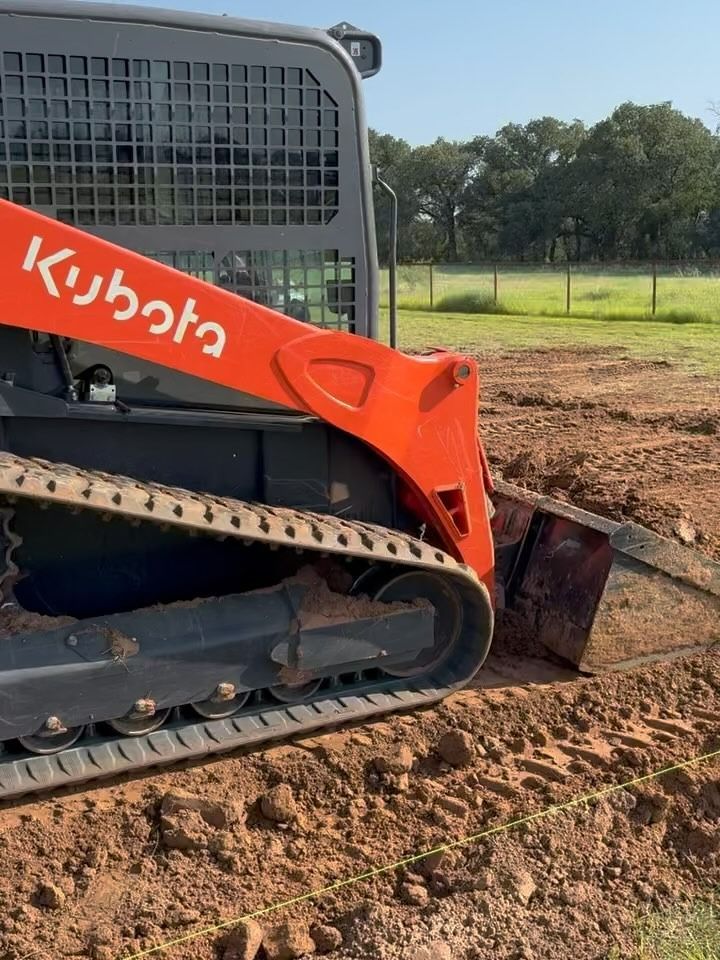 Orange Kubota track loader digging a trench in dirt, outdoors.