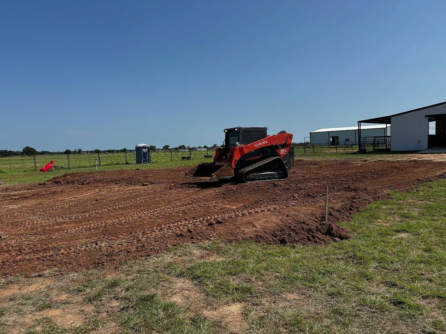 Red skid steer grading a dirt area in a grassy field. Building and blue portable toilet visible.