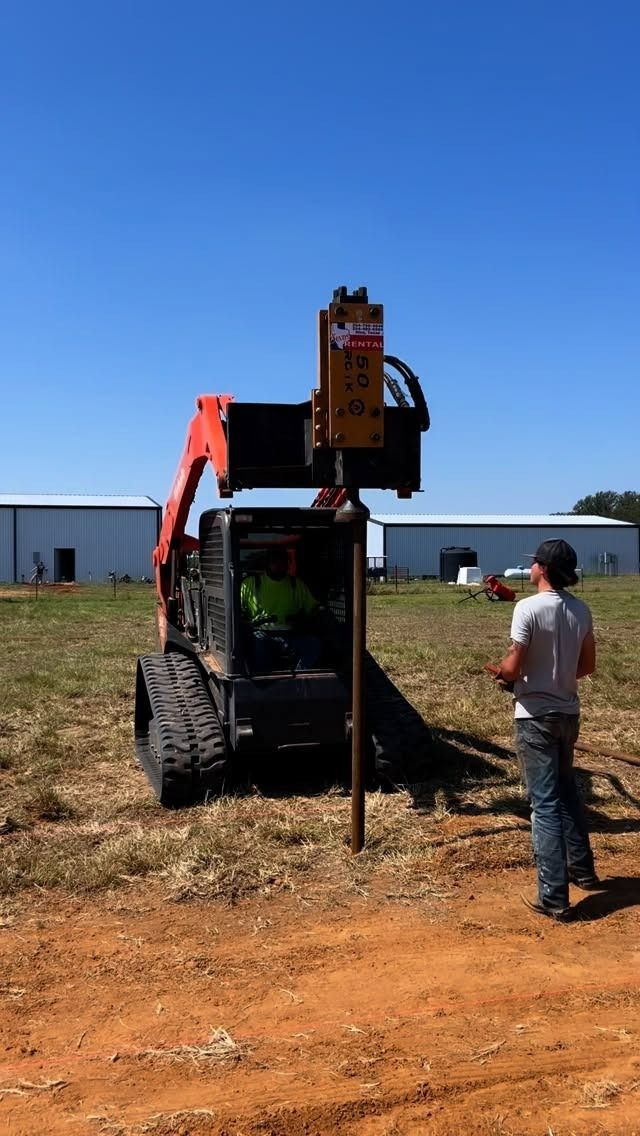 Skid steer using a post driver to install a metal post; worker observes. Outdoors, sunny day.