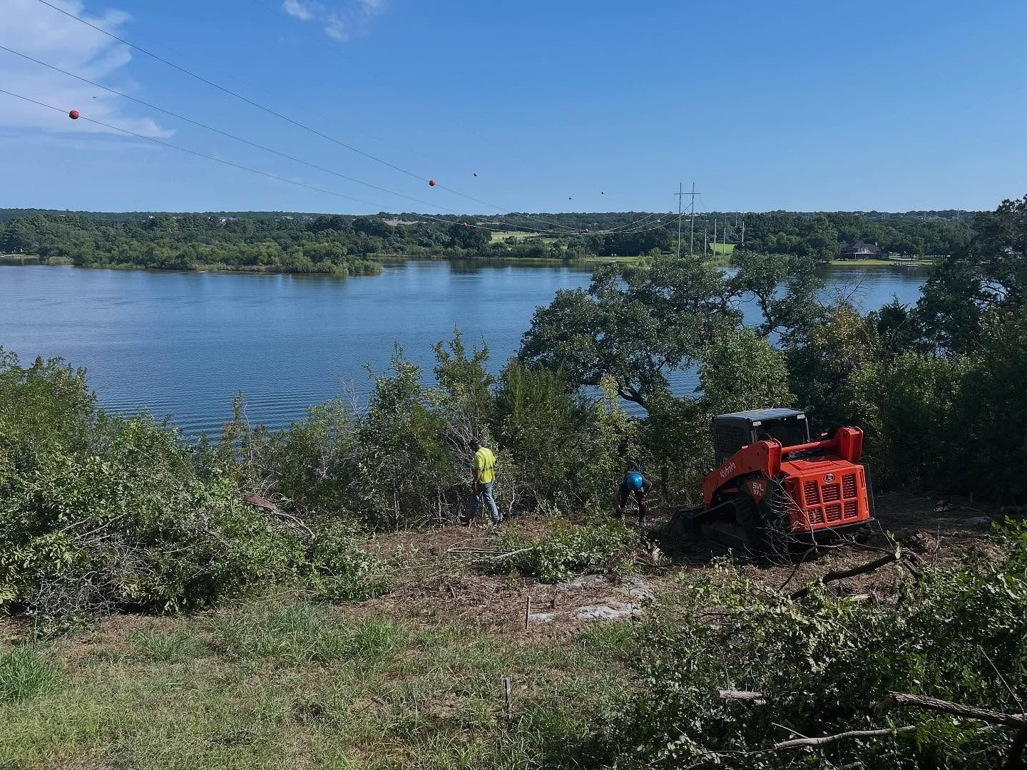 Construction workers and orange tractor clearing brush near a lake on a sunny day.