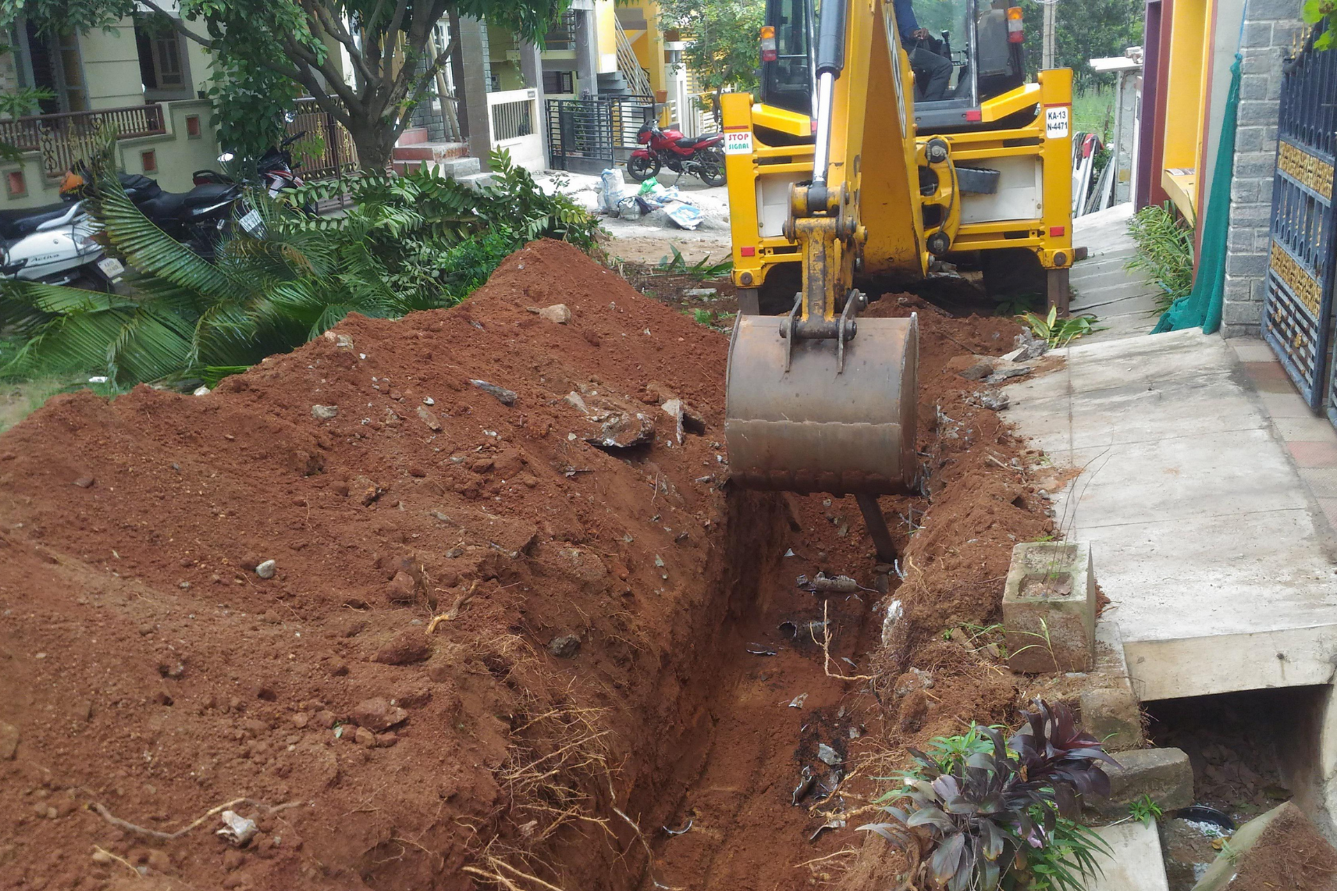 Yellow backhoe digging a trench in red soil next to a concrete walkway.