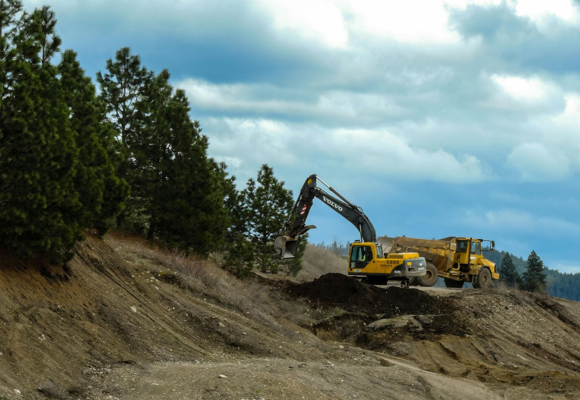 An excavator and dump truck on a hillside under a cloudy sky, removing earth near evergreen trees.