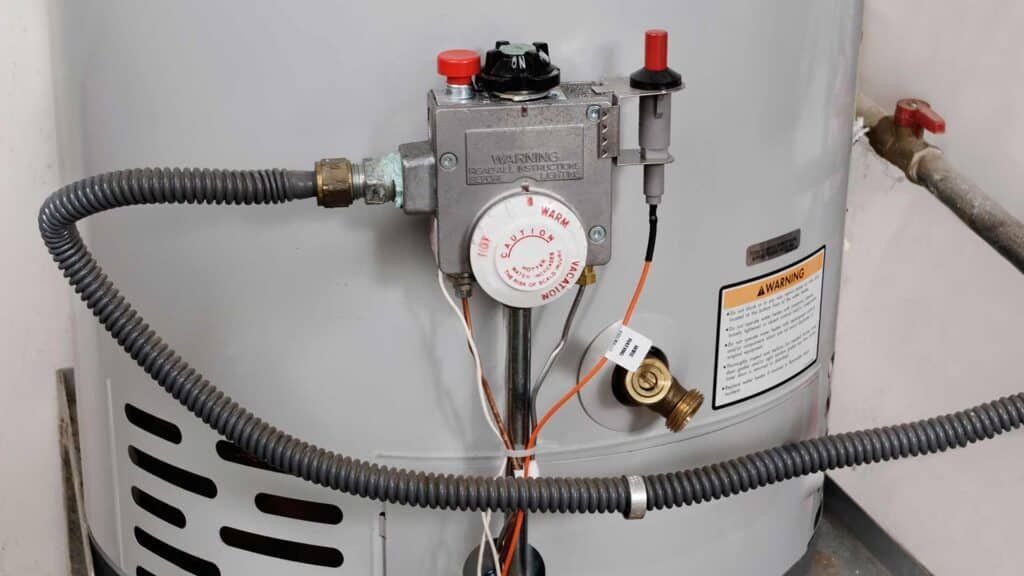 Gas water heater with control valve, corrugated hose, and warning labels against a wall.