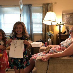 A little girl is holding a piece of paper in front of a woman sitting on a couch.