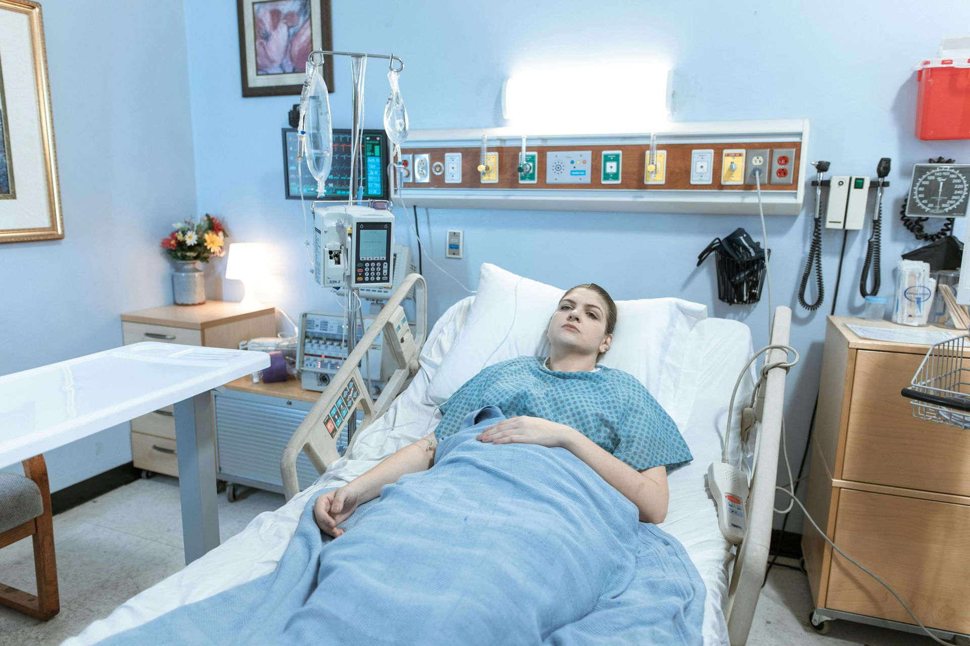 Woman in hospital bed, wearing gown, with IV drip, in a pale blue room.